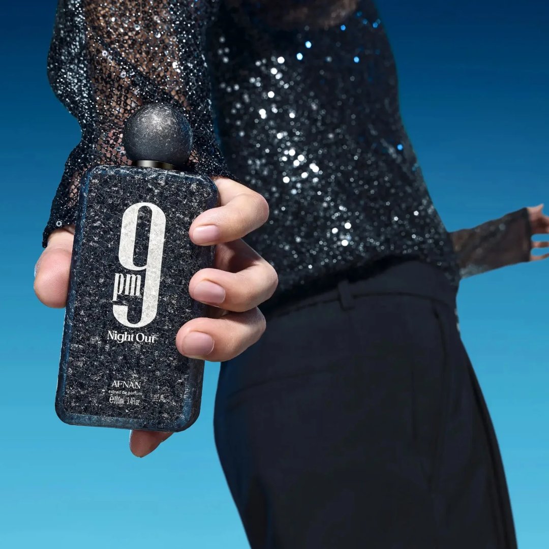 Afnan 9 PM Night Out Extrait de Parfum 100ml — A person in a sparkly black top holds a dark, textured perfume bottle with '9 pm Night Out' visible on the front against a bright blue sky background. | Afnan 9 PM Night Out Extrait de Parfum 100ml — Seseorang dalam atasan hitam berkilauan memegang botol minyak wangi