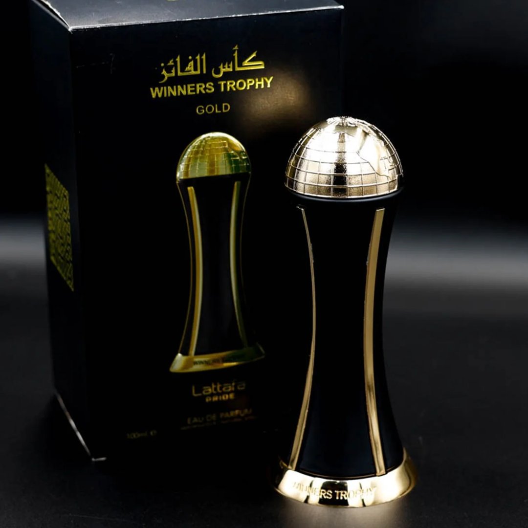 Lattafa Pride Al Khas Winners Trophy Gold 100ml — A black box with gold Arabic and English text sits next to a matching black and gold trophy-shaped perfume bottle with a golden globe top, set against a dark background. | Lattafa Pride Al Khas Winners Trophy Gold 100ml — Kotak hitam dengan teks emas Arab dan Inggeris