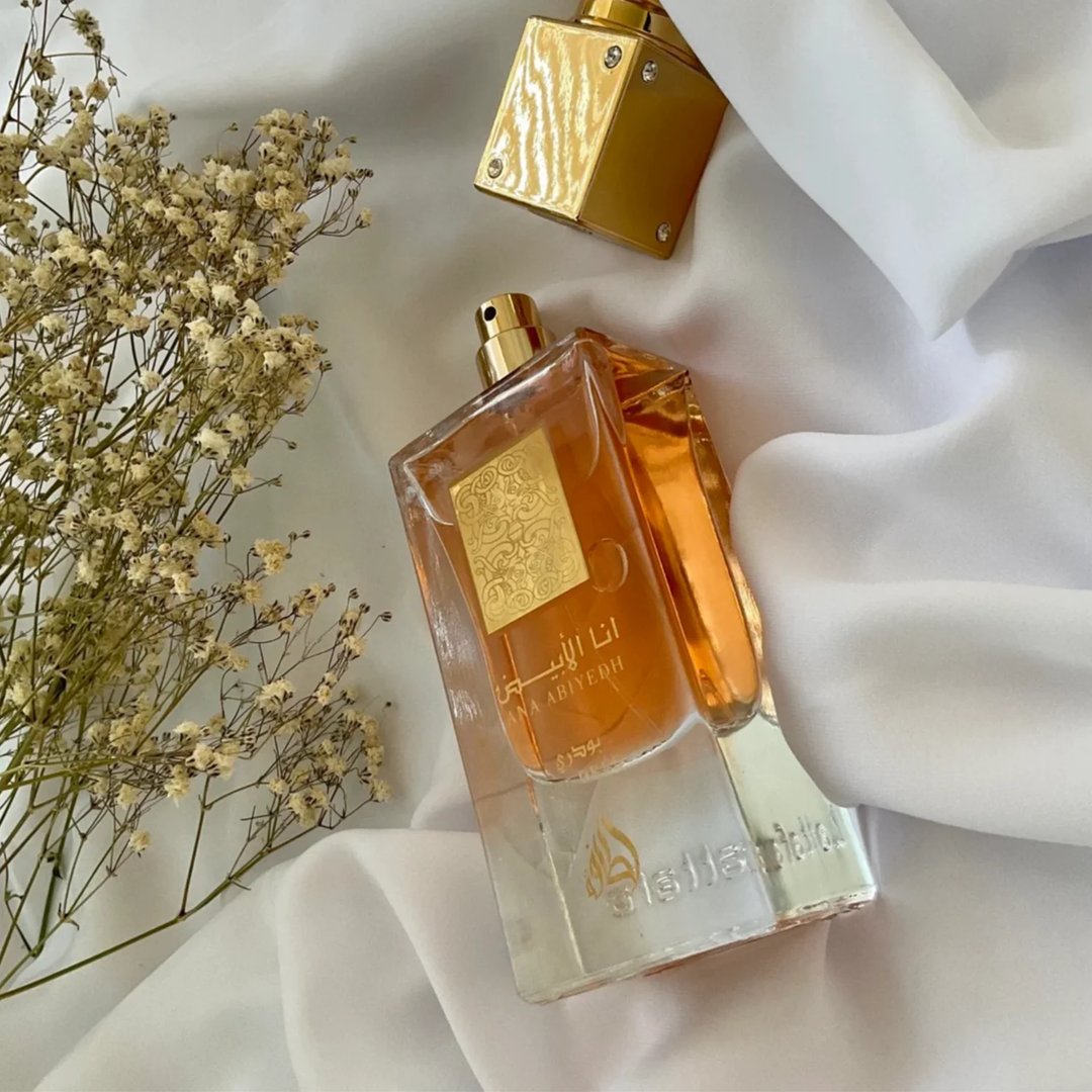 Lattafa Ana Abiyedh Poudree by Lattafa 60ml — Amber liquid in a clear glass bottle with a gold label, next to dried white flowers on a white satin cloth. Another gold bottle is in the background. | Lattafa Ana Abiyedh Poudree by Lattafa 60ml — Cecair amber dalam botol kaca jernih dengan label emas, bersebelahan bunga