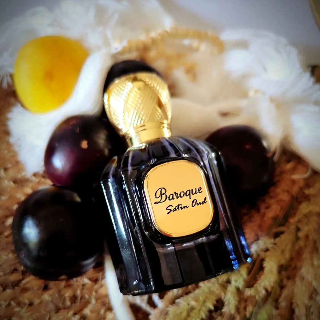 Maison Alhambra Baroque Satin Oud 100ml — A dark glass perfume bottle with a gold fluted cap and a gold label reading "Baroque Satin Oud" sits among dark purple fruits, a yellow fruit, and white feathers on a textured background. | Maison Alhambra Baroque Satin Oud 100ml — Botol minyak wangi kaca gelap dengan penutup