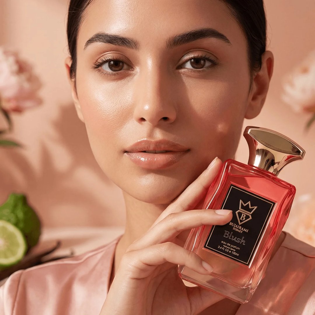 Bujairami Blush by Bujairami Eau de Parfum 100ml — Woman holding a pink glass perfume bottle with a gold cap, with blurred flowers and lime in the background.