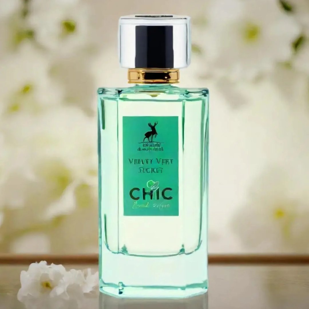 Maison Alhambra Chic Velvet Vert Secret 100ml — A clear glass perfume bottle with green liquid and a green label featuring a deer logo and text, set against a soft, blurred background of white flowers. | Maison Alhambra Chic Velvet Vert Secret 100ml — Botol kaca jernih berisi cecair hijau dan label hijau menampilkan