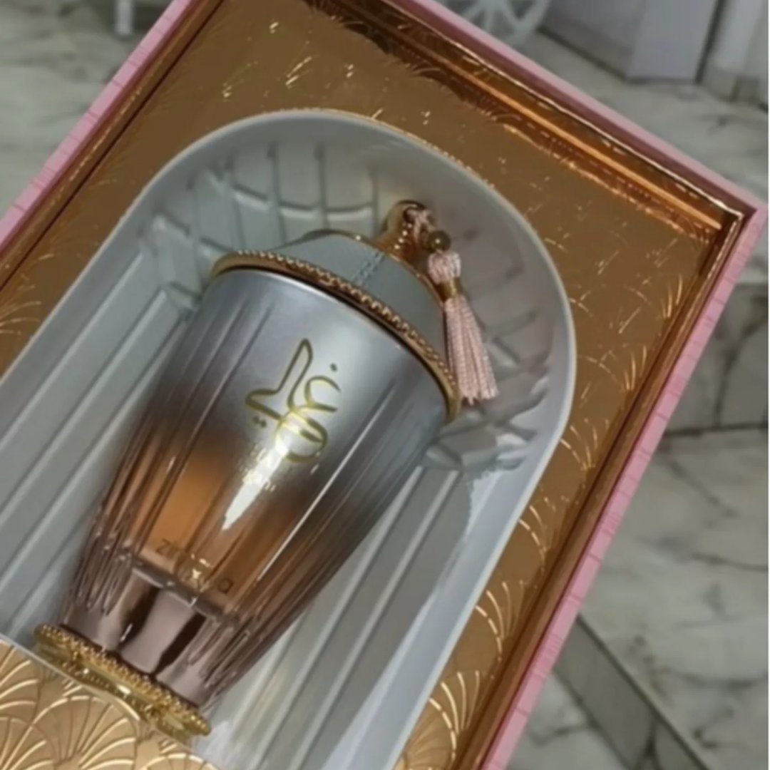 Zimaya Ghali Elegant Eau De Parfum 100ml by Zimaya — A silver and rose gold fluted perfume bottle with Arabic calligraphy and a pink tassel sits in a pink and gold patterned box. | Zimaya Ghali Elegant Eau De Parfum 100ml by Zimaya — Botol minyak wangi perak dan emas mawar berlekuk dengan kaligrafi Arab dan juntai