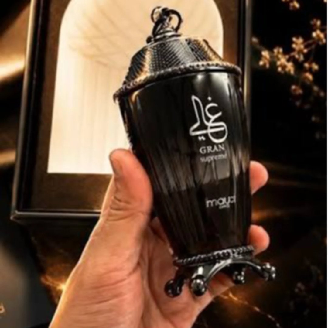 Zimaya Ghali Imperial Eau De Parfum 100ml by Zimaya — A hand holds a black, ornate perfume bottle with gold Arabic calligraphy and "GRAN SUPREME" text, set against a blurred background with a golden archway and branches. | Zimaya Ghali Imperial Eau De Parfum 100ml by Zimaya — Tangan memegang botol minyak wangi hitam