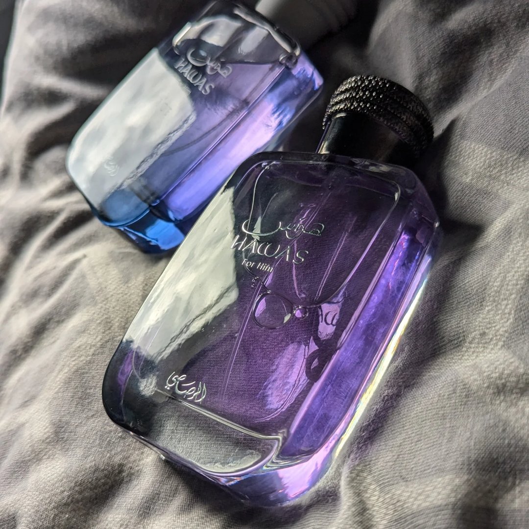 Rasasi Hawas For Him Eau De Parfum — Two glass perfume bottles with purple and blue gradients, one with a textured black cap, resting on a grey textured fabric surface. | Dua botol minyak wangi kaca dengan gradien ungu dan biru, satu dengan penutup hitam bertekstur, diletakkan di atas permukaan fabrik kelabu