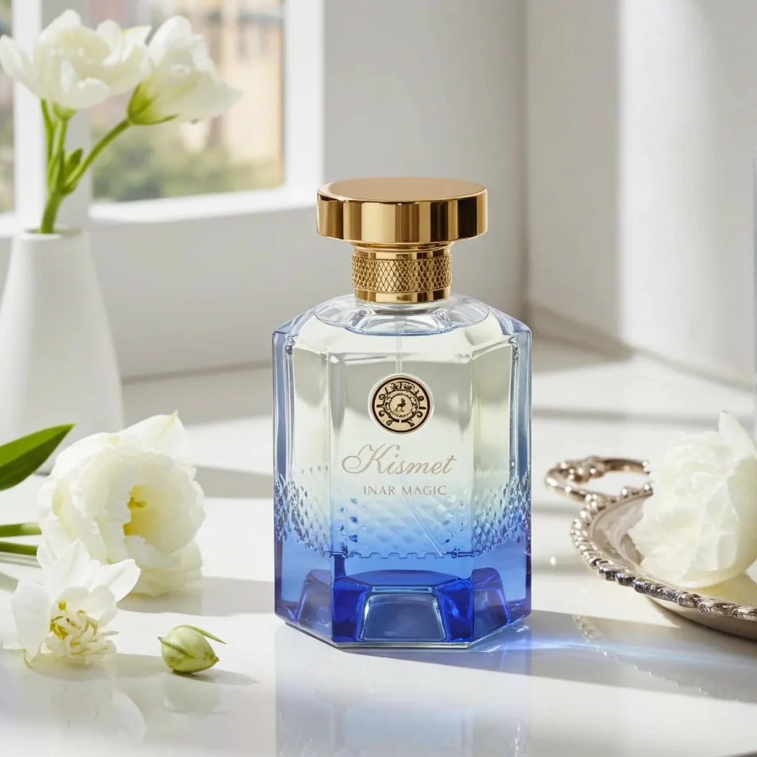 Maison Alhambra Kismet Lunar Magic 100ml by Maison Alhambra — A faceted glass perfume bottle with a gold cap and blue gradient base, surrounded by white flowers and a decorative silver tray on a white surface. | Maison Alhambra Kismet Lunar Magic 100ml by Maison Alhambra — Botol minyak wangi kaca berfaset dengan