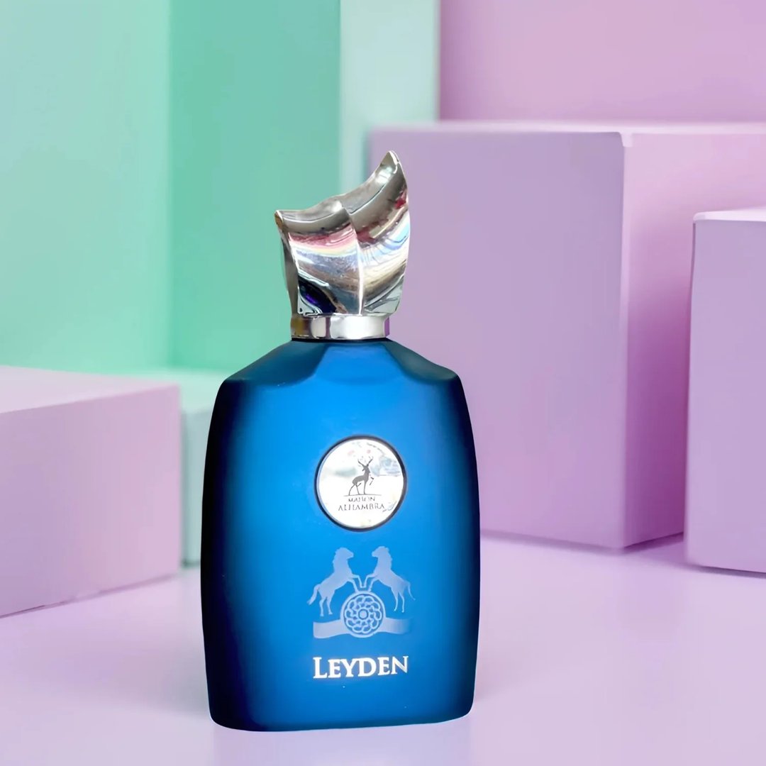 Maison Alhambra Leyden by Maison Alhambra Eau De Parfum 100ml — A blue glass perfume bottle with a silver cap and embossed emblem, set against a pastel background with geometric shapes. | Maison Alhambra Leyden by Maison Alhambra Eau De Parfum 100ml — Botol kaca biru dengan penutup perak dan lambang timbul, berlatar