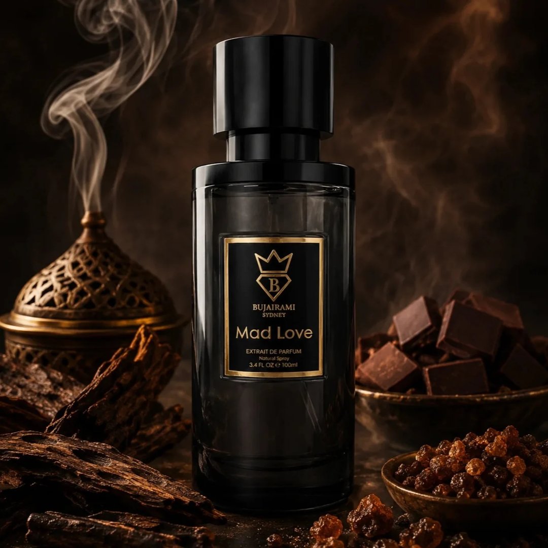 Bujairami Mad Love 100ml Extrait Unisex — Dark glass perfume bottle with a black cap, gold label featuring a crown and 'B' logo, surrounded by dark wood pieces, chocolate cubes, and amber-colored crystals.