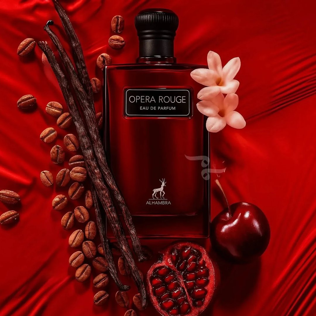 Maison Alhambra Opera Rouge — A red perfume bottle is surrounded by coffee beans, vanilla pods, a halved pomegranate, a cherry, and two light pink flowers on a red satin background. | Maison Alhambra Opera Rouge — Botol minyak wangi merah dikelilingi oleh biji kopi, batang vanila, sebiji delima yang dibelah dua
