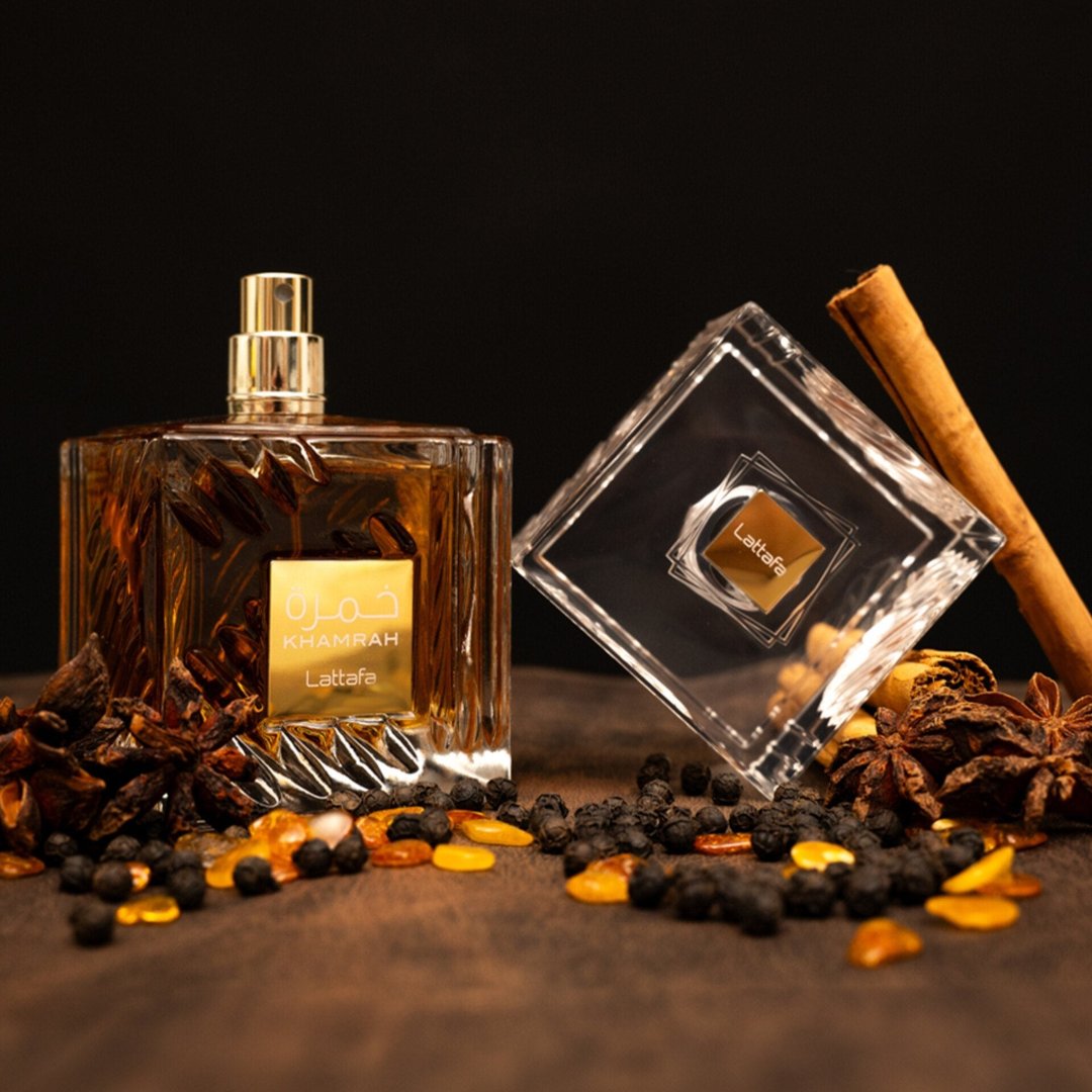Lattafa Khamrah by Lattafa 100ml EDP Unisex — Amber liquid perfume bottle with gold cap, tilted clear square cap, cinnamon stick, star anise, peppercorns, and amber pieces on a wooden surface with dark background.