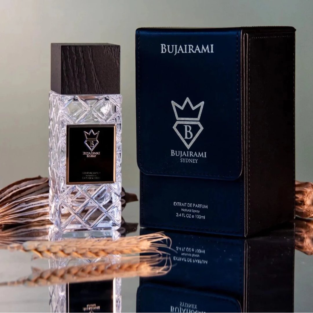 Bujairami Shadow Script - Bujairami 100ml Extrait, clear faceted glass bottle with dark cap, next to navy blue presentation box with silver logo, on reflective surface with dried wheat and seed pods.