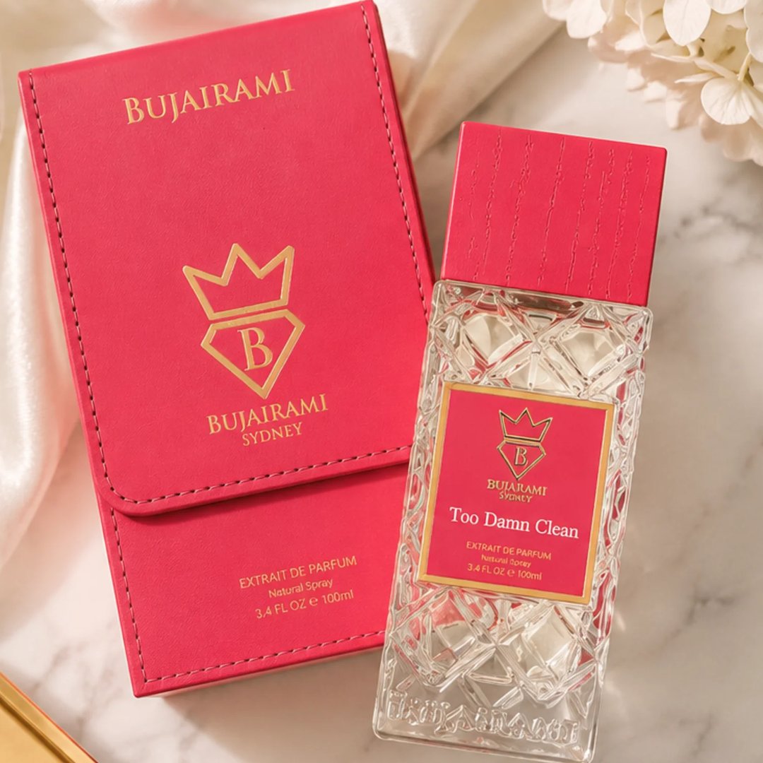 Bujairami Too Damn Clean — A clear glass perfume bottle with a textured red cap and a pink label featuring a gold crown and 'B' logo, placed next to its matching pink presentation box with gold lettering and logo.