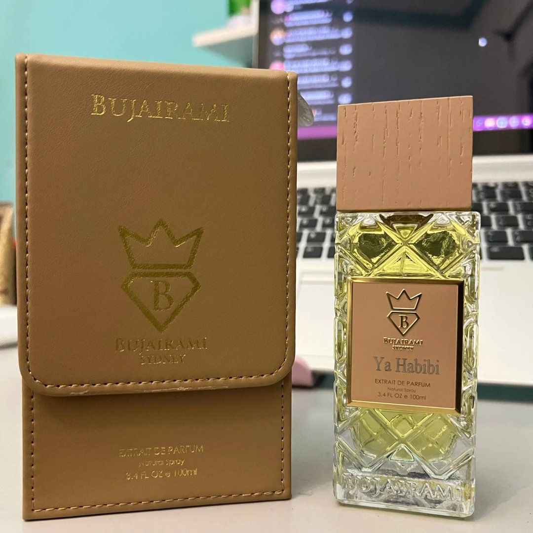 Bujairami Ya Habibi — Gold-embossed brown leather case next to a clear glass perfume bottle with a textured wood-like cap and gold label.