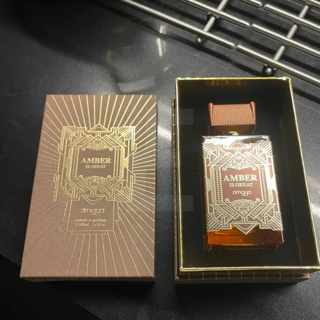 Zimaya Amber Is Great Eau De Parfum 100ml — A brown textured box with gold art deco design is open next to a gold-lined box containing a glass perfume bottle with a similar gold art deco design and a brown ribbed cap. | Zimaya Amber Is Great Eau De Parfum 100ml — Kotak bertekstur coklat dengan reka bentuk art deco