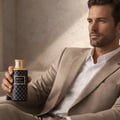 Maison Asrar Legacy by Maison Asrar 100ml EDP Unisex — Lelaki berbaju beige pegang botol minyak wangi corak biru gelap dan emas dengan penutup emas