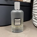 Khadlaj Magnate Noble by Khadlaj 100ml EDP — A frosted grey perfume bottle with a textured silver cap sits on a textured surface, with a black cabinet and striped fabric in the background. | Khadlaj Magnate Noble by Khadlaj 100ml EDP — Botol minyak wangi kelabu beku dengan penutup perak bertekstur diletakkan di atas