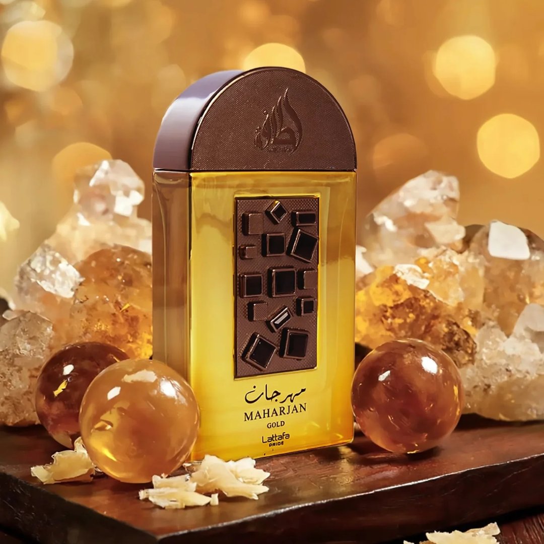 Lattafa Pride Maharjan Gold — A golden perfume bottle with a textured brown cap and a chocolate-like decorative panel, surrounded by amber-colored crystals and spheres on a dark wooden surface, with a bokeh background of golden lights. | Lattafa Pride Maharjan Gold — Botol minyak wangi keemasan dengan penutup coklat