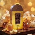Lattafa Pride Maharjan Gold — A golden perfume bottle with a textured brown cap and a chocolate-like decorative panel, surrounded by amber-colored crystals and spheres on a dark wooden surface, with a bokeh background of golden lights. | Lattafa Pride Maharjan Gold — Botol minyak wangi keemasan dengan penutup coklat
