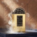 Lattafa Pride Maharjan Gold — A gold perfume bottle with black geometric accents, surrounded by wisps of smoke against a textured brown background. | Lattafa Pride Maharjan Gold — Botol minyak wangi emas dengan aksen geometri hitam, dikelilingi oleh kepulan asap berlatarkan latar belakang coklat bertekstur.