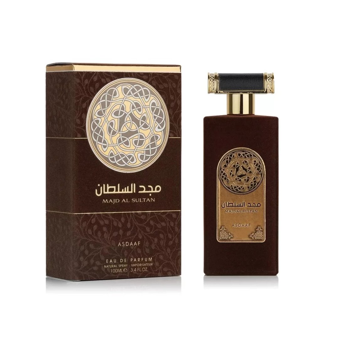 Asdaaf Majd Al Sultan by Asdaaf 100ml EDP — A brown velvet perfume bottle and its matching box are displayed side-by-side against a white background. Both feature a circular gold and white Celtic knot design and Arabic text. | Asdaaf Majd Al Sultan by Asdaaf 100ml EDP — Sebotol minyak wangi baldu coklat dan kotaknya