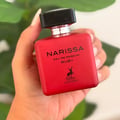 Maison Alhambra Narissa Ruby — A hand holds a red square perfume bottle with a black cap and silver accents. The bottle has black text reading 'NARISSA EAU DE PARFUM RUBY' and a deer silhouette logo. | Maison Alhambra Narissa Ruby — Tangan memegang botol pewangi merah berbentuk segi empat dengan penutup hitam dan