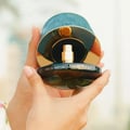 Maison Asrar Never Forget Me by Maison Asrar 90ml EDP Women — Tangan memegang botol minyak wangi kaca gelap dengan penutup bertekstur teal dan aksen emas, menunjukkan label 'EAU DE PARFUM Never Forget Me'.