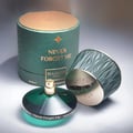 Maison Asrar Never Forget Me oleh Maison Asrar 90ml EDP Wanita — Kotak silinder hijau bertekstur dengan penutup emas, botol minyak wangi terbuka dengan kaca hijau dan muncung semburan emas, serta penutup bertekstur berombak yang condong.
