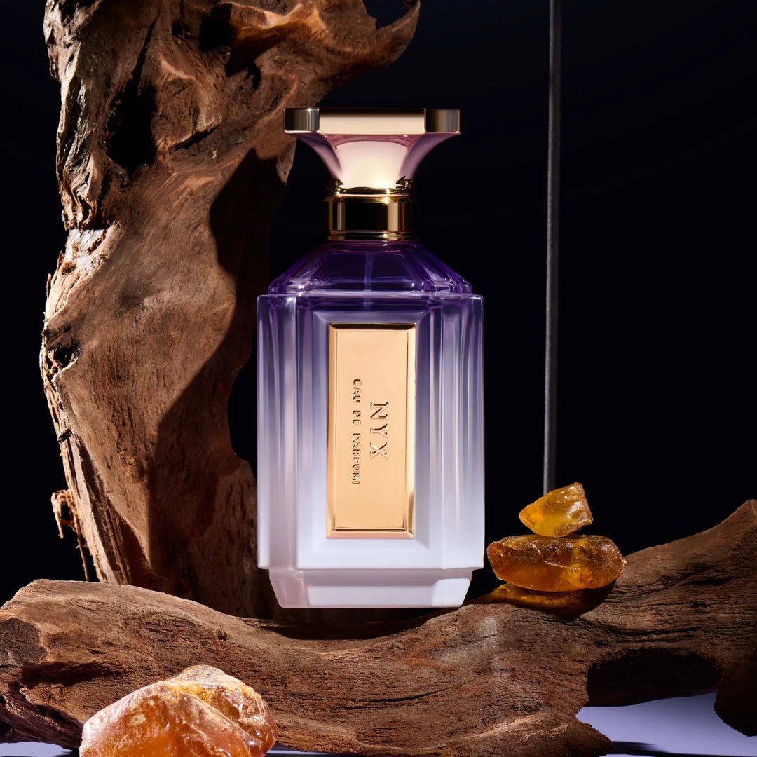 Velixir Nyx Velixir Parfums 100ml for Unisex — A faceted purple and white glass perfume bottle with a gold cap and label, placed among rough wood and amber-like stones on a dark background. | Velixir Nyx Velixir Parfums 100ml for Unisex — Botol kaca ungu dan putih berfaset dengan penutup dan label emas, diletakkan di