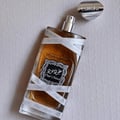 Lattafa Oud Mood Reminiscence Lattafa 100ml — A clear glass perfume bottle with amber liquid, wrapped in white ribbon with repeating text, lies on a textured white surface with the silver cap placed nearby. | Lattafa Oud Mood Reminiscence Lattafa 100ml — Botol minyak wangi kaca jernih dengan cecair berwarna ambar