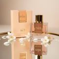 Lattafa Ana Abiyedh Coral — A coral-colored perfume bottle and its box sit on a pink, sheer fabric with pearls and small