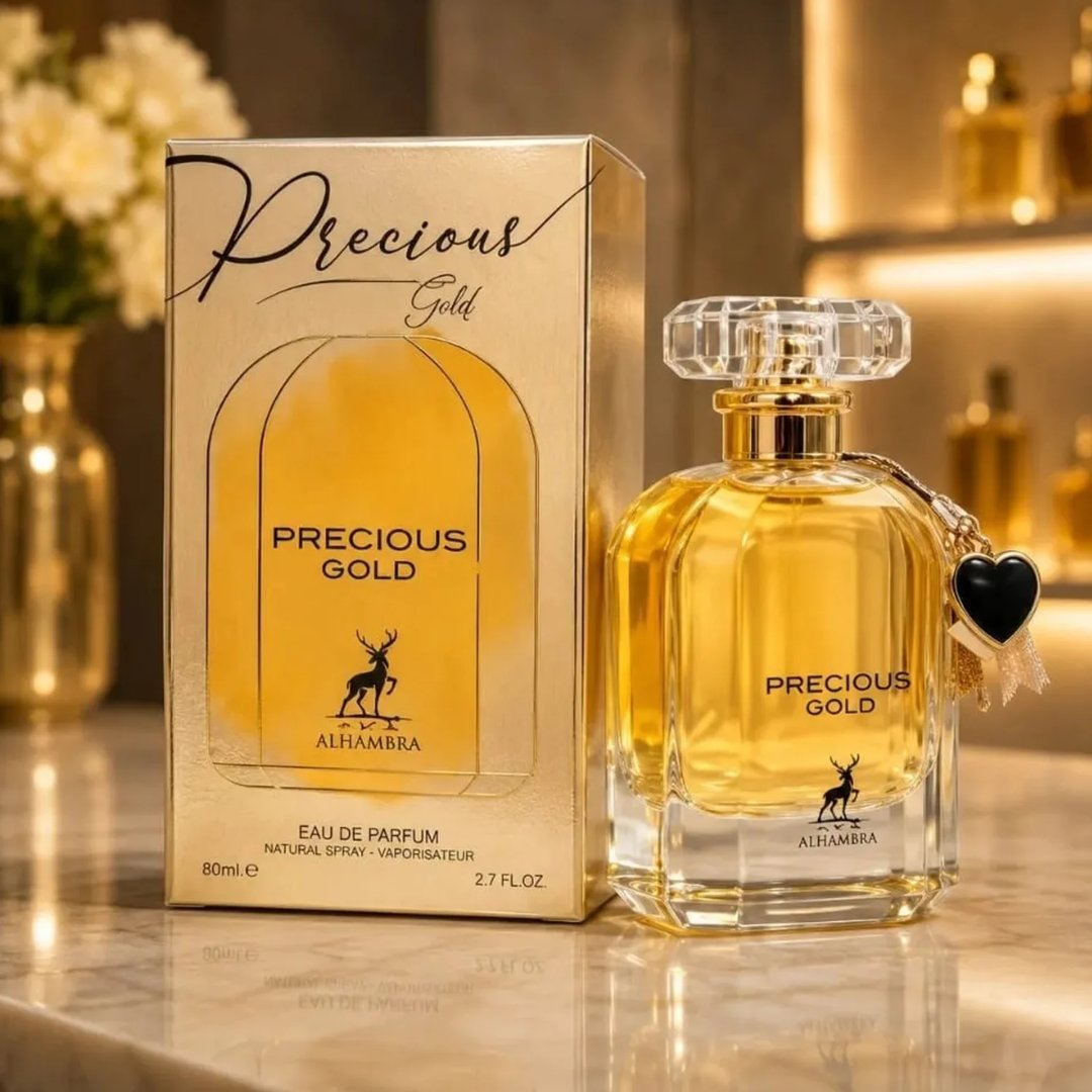 Maison Alhambra Precious Gold 80ml — A gold-toned perfume bottle with a faceted crystal cap and a black heart charm sits next to its matching gold box, with a vase of white flowers in the background. | Maison Alhambra Precious Gold 80ml — Botol minyak wangi berwarna emas dengan penutup kristal berfaset dan loket hati
