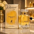 Maison Alhambra Precious Gold 80ml — A gold-toned perfume bottle with a faceted crystal cap and a black heart charm sits next to its matching gold box, with a vase of white flowers in the background. | Maison Alhambra Precious Gold 80ml — Botol minyak wangi berwarna emas dengan penutup kristal berfaset dan loket hati