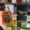 Ard Al Zaafaran Qaed Al Fursan — A hand holds a clear glass perfume bottle with amber liquid and a gold cap, next to its black and gold box featuring a running horse. | Ard Al Zaafaran Qaed Al Fursan — Tangan memegang botol minyak wangi kaca jernih dengan cecair ambar dan penutup emas, bersebelahan kotak hitam dan