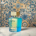 Lattafa Sheikh Al Shuyukh Supreme — A turquoise glass perfume bottle with a textured gold cap sits on a marble surface against a blue and gold patterned backdrop. | Lattafa Sheikh Al Shuyukh Supreme — Botol kaca berwarna biru kehijauan dengan penutup emas bertekstur diletakkan di atas permukaan marmar berlatarkan