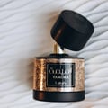 Lattafa Tamima by Lattafa 100 ml Eau de Parfum — A black and gold perfume bottle with an ornate textured gold band and a tilted wooden cap, resting on a white textured surface. | Lattafa Tamima by Lattafa 100 ml Eau de Parfum — Botol minyak wangi hitam dan emas dengan jalur emas bertekstur berukir dan penutup kayu