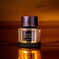 Lattafa Tamima — A dark glass perfume bottle with ornate gold detailing and a black cap sits on a reflective surface against a warm, blurred background. | Botol minyak wangi kaca gelap dengan perincian emas berornamen dan penutup hitam duduk di permukaan reflektif berlatarkan latar belakang yang hangat dan kabur.