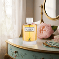 Maison Alhambra La Vivacite by Maison Alhambra 100ml EDP — Amber perfume bottle with white cap and pink ribbon, placed on a vintage vanity table with a pink rose and lace curtains.