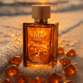 Maison Alhambra Your Touch Amber — Amber glass perfume bottle with gold cap, surrounded by amber-colored stones on a wet sandy beach at sunset.