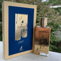 Maison Alhambra Minerale Gold Eau de Parfum — Gold bottle and box with blue and gold abstract design, set against a blurred green outdoor background.