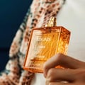 Rayhaan Adonis Icarus Rayhaan Perfume 100ml — A hand holds an amber-colored glass perfume bottle with a gold cap, featuring embossed patterns and white text. The background shows a blurred patterned scarf and dark blue fabric. | Rayhaan Adonis Icarus Rayhaan Perfume 100ml — Tangan memegang botol minyak wangi kaca