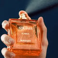 Rayhaan Adonis Icarus Rayhaan Perfume 100ml — A hand sprays the orange glass perfume bottle against a dark blue background. | Rayhaan Adonis Icarus Rayhaan Perfume 100ml — Tangan menyembur botol minyak wangi kaca berwarna oren berlatarkan latar biru gelap.