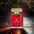 Al Wataniah Rawaee Monarch Eau De Parfum 100ml — A red glass perfume bottle with a gold cap sits on a reflective surface, surrounded by sparkling gold lights against a dark background. | Al Wataniah Rawaee Monarch Eau De Parfum 100ml — Botol kaca merah untuk minyak wangi dengan penutup emas terletak di atas permukaan