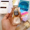 Lattafa Ana Abiyedh Leather 60ml by Lattafa Perfumes — A hand holds a clear glass perfume bottle with a gold cap and ornate label, surrounded by other perfume boxes and bottles. | Lattafa Ana Abiyedh Leather 60ml by Lattafa Perfumes — Tangan memegang botol minyak wangi kaca jernih dengan penutup emas dan label