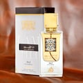Lattafa Ana Abiyedh Leather 60ml by Lattafa Perfumes — A clear glass perfume bottle with a gold cap and ornate black and gold label, standing next to its white and gold patterned box on a textured brown background. | Lattafa Ana Abiyedh Leather 60ml by Lattafa Perfumes — Botol minyak wangi kaca jernih bertutup emas