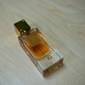 Lattafa Ana Abiyedh Poudree by Lattafa 60ml — A clear glass perfume bottle with amber liquid and a gold cap, resting on a light wood grain surface. The bottle has an ornate gold label and Arabic script. | Lattafa Ana Abiyedh Poudree by Lattafa 60ml — Botol kaca jernih berisi cecair ambar dan penutup emas, diletakkan