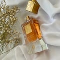 Lattafa Ana Abiyedh Poudree by Lattafa 60ml — Amber liquid in a clear glass bottle with a gold label, next to dried white flowers on a white satin cloth. Another gold bottle is in the background. | Lattafa Ana Abiyedh Poudree by Lattafa 60ml — Cecair amber dalam botol kaca jernih dengan label emas, bersebelahan bunga