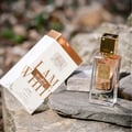 Lattafa Ana Abiyedh Poudree 60ml — A white and gold perfume box tilted on its side next to a clear glass perfume bottle with a gold cap, both resting on grey rocks with a blurred natural background. | Lattafa Ana Abiyedh Poudree 60ml — Kotak minyak wangi putih dan emas diletakkan senget di sebelah botol minyak wangi