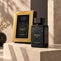 Lattafa Bade'e Al Oud Black Exposed Lattafa 100ml — Black perfume bottle and its gold-trimmed gift box displayed on textured stone surfaces, with a blurred dried plant in the background.
