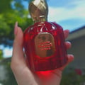 Maison Alhambra Baroque Rouge Extrait 100ml by Maison Alhambra — A hand holds a red, faceted glass perfume bottle with a gold cap, outdoors with blurred green foliage and blue sky in the background. | Maison Alhambra Baroque Rouge Extrait 100ml by Maison Alhambra — Tangan memegang botol minyak wangi kaca merah