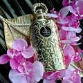 Lattafa Bayaan Eau De Parfum 100ml by Lattafa — A gold-embossed perfume bottle rests on a golden leaf-shaped tray, surrounded by delicate pink orchids against a dark, ribbed background. | Lattafa Bayaan Eau De Parfum 100ml by Lattafa — Botol minyak wangi bersalut emas diletakkan di atas dulang berbentuk daun emas